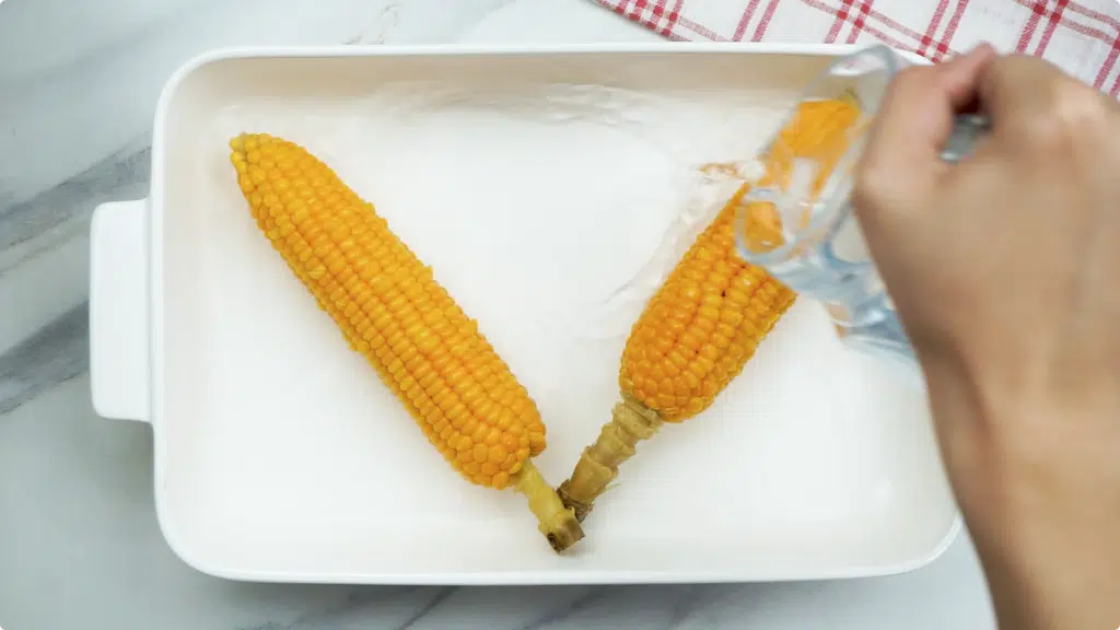 Two corn on the cob in a white baking dish being covered with water