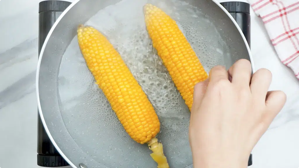 A lady's hands placing two corn on the cob into a skillet with simmering water