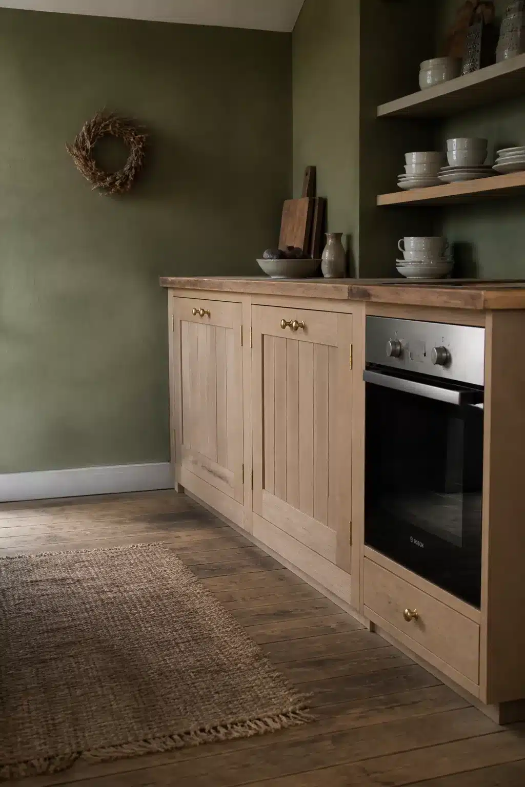 Moss green kitchen walls with bare oak shaker cabinets, brass knobs, open shelves, and jute rug