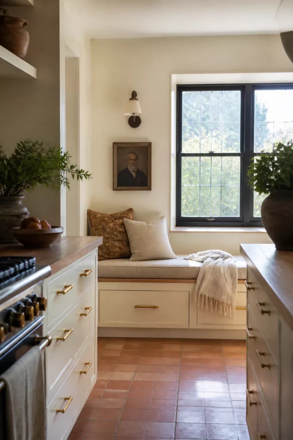 Cream kitchen with brass handles, terracotta tile floor, and built-in window seat with black-framed window