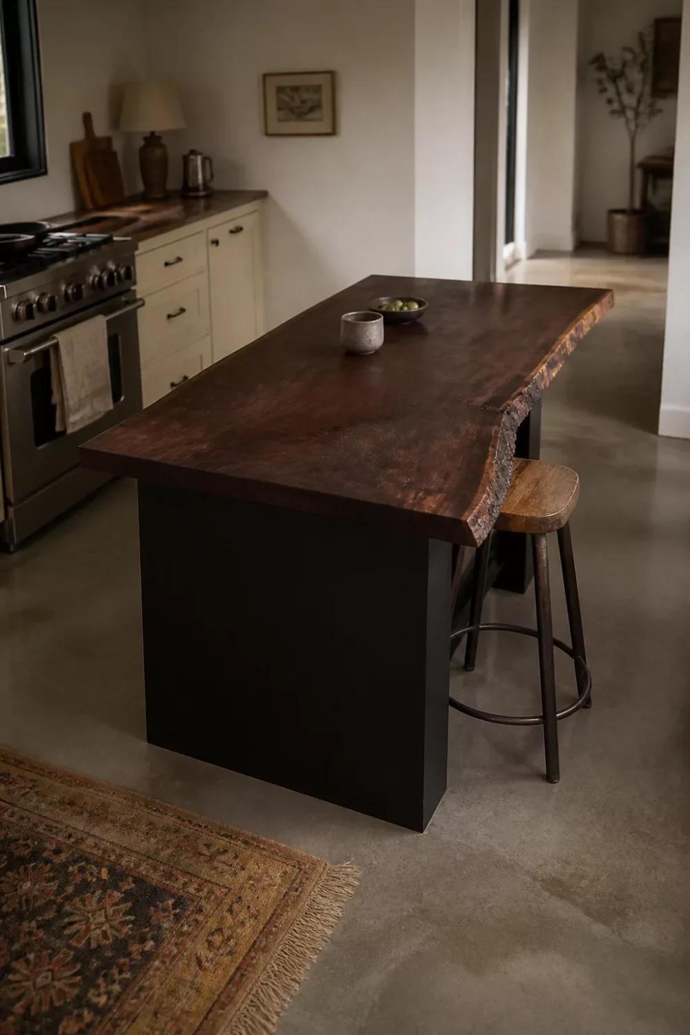 Live-edge dark wood kitchen countertop on a matte black island base with concrete floors and a cream range kitchen.