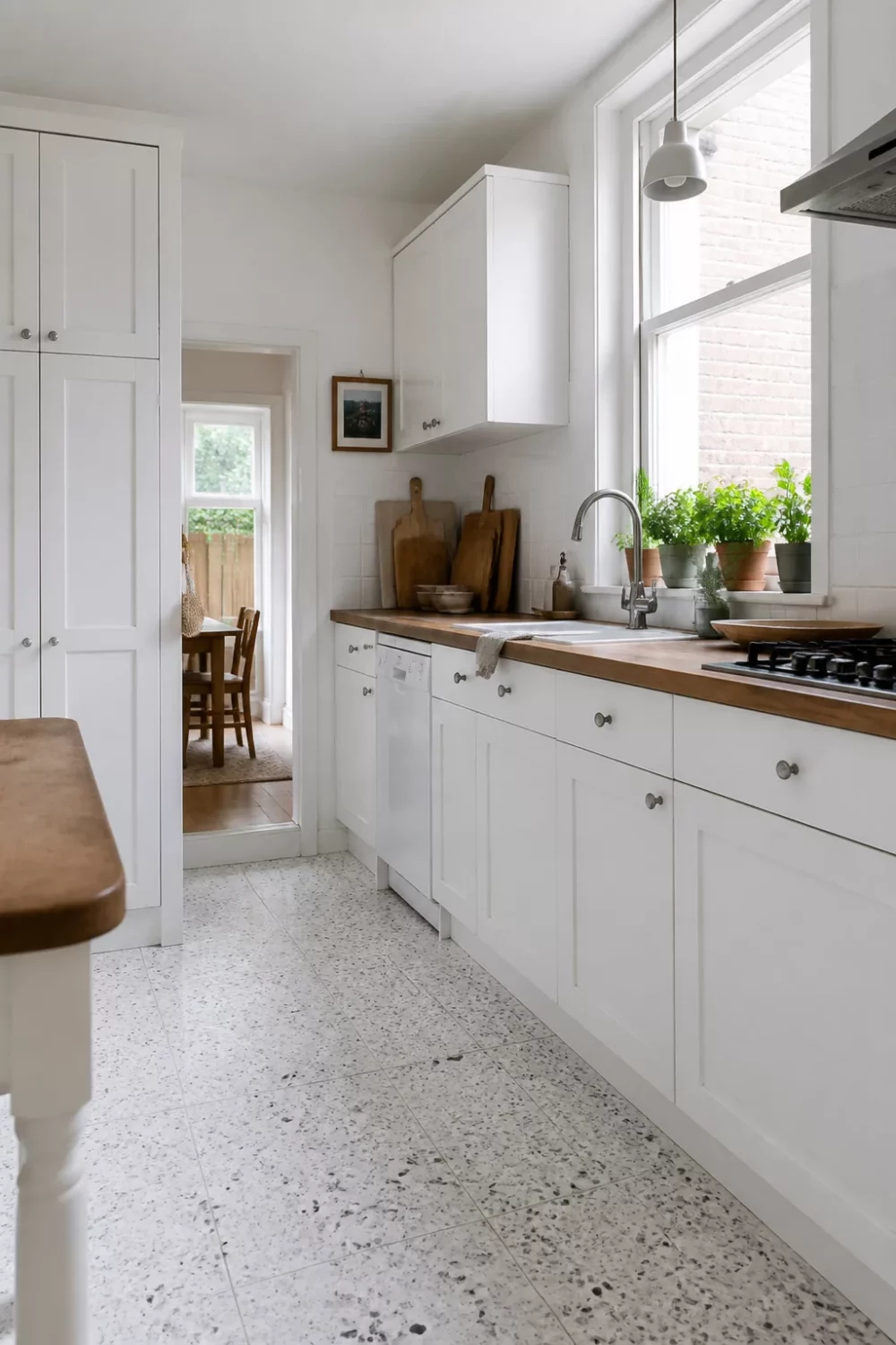 White kitchen with wood countertops, Shaker cabinets, and terrazzo floor tiles — galley layout with herb-lined window